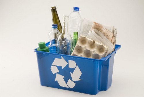 A blue recycling bin positioned against a plain light background, filled with various types of waste materials. Inside the bin, there are several clear and colored plastic bottles, including green, blue, and transparent ones, with some caps visible. Additionally, there are brown paper egg cartons, crumpled sheets of newspaper or paper waste, and an empty glass bottle leaning against the side of the bin. The bin features a white recycling symbol on its side, indicating it is intended for recyclable rubbish. The waste items appear loosely stacked, with some objects extending above the rim of the bin. The environment suggests an indoor setting or a designated waste collection point, common in urban or suburban areas such as Surbiton, where professional rubbish collection services like Office Clearance Surbiton operate to handle waste removal and recycling. The overall scene emphasizes the sorting of recyclable waste materials, showcasing the typical contents of a bin used for household or small-scale commercial waste disposal, aligned with environmentally responsible rubbish management practices in the UK.