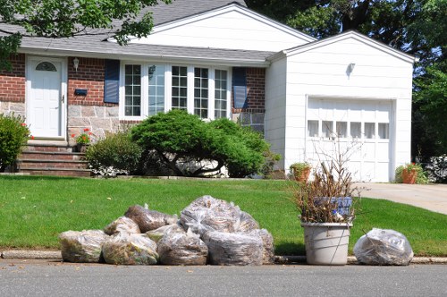 In front of a residential property, a collection of irregularly shaped rocks and boulders with rough, weathered surfaces are placed on the curbside in a suburban neighbourhood, alongside a metal bucket containing dried plants and twigs. The house in the background features a brick façade with dark blue shutters, a white front door, and large bay windows, with a well-maintained green lawn and neatly trimmed shrubbery. A paved driveway runs up to an attached garage with a white door, and a stone pathway with steps leads to the entrance. The scene appears to depict a clear day, with natural sunlight illuminating the outdoor setting. The image relates to rubbish removal services in Surbiton, focusing on the collection of garden waste and loose debris for clearance, managed by Office Clearance Surbiton, situated in the KT town postcode area.
