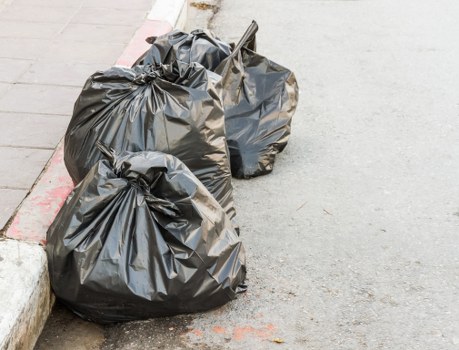 Two large black rubbish bags made of thick plastic material, slightly wrinkled and tied at the top, are placed on the edge of a paved street or driveway next to a curb with a red marking. The bags contain mixed waste, possibly domestic or office refuse, and are positioned close to the grey concrete pavement, with a small metal tool or pole leaning against one of the bags. The background shows a smooth asphalt surface, indicating an outdoor urban environment typical of Surbiton, with no additional objects or vehicles visible. The scene suggests a scheduled rubbish collection or a waste clearance process undertaken by Office Clearance Surbiton as part of local rubbish removal services, with the bags arranged for disposal near residential or commercial premises within the Surbiton area, postcode KT6.
