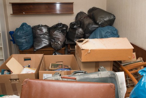 The image shows a collection of black and blue rubbish bags, some tightly sealed and others slightly bulging, positioned on a wooden bench against a beige wall inside an indoor space, possibly a storage or disposal area. Several cardboard boxes are visible in the foreground; one is partially open revealing newspapers and miscellaneous paper waste, while another appears to contain small items or packaging. A large, silver metal object, likely a ventilation duct or similar industrial component, is lying on the floor near the boxes. To the right, part of a wooden drying rack or small shelving unit is visible, with blue plastic bags hanging or placed on it. The background includes a wooden shelf mounted on the wall above the rubbish bags, and a section of what appears to be a wooden door or panel to the far right. The area appears cluttered, with items arranged haphazardly, indicating an active rubbish or clearance activity commonly associated with waste removal services in the Surbiton area.