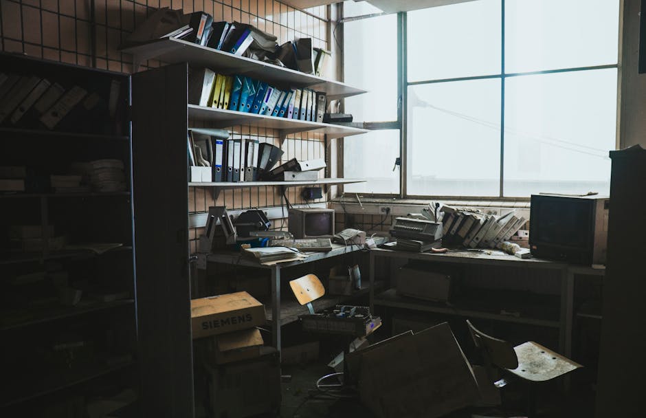 A light-colored wooden table in a workspace or studio environment is cluttered with various discarded items, including two cardboard boxes—one larger with rectangular cut-out handles on its sides an