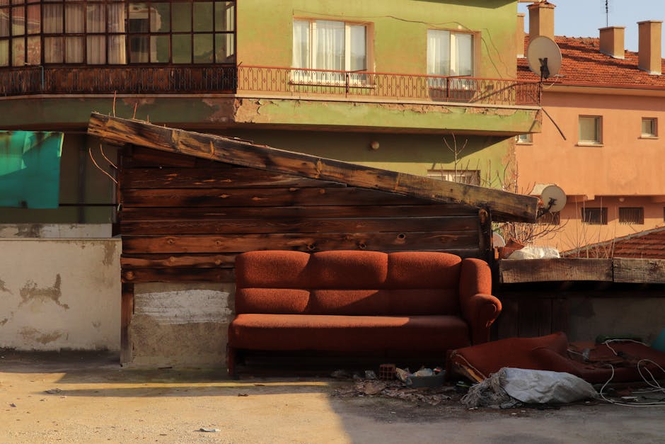 A vintage-style, upholstered sofa with a wood frame and intricate carvings is positioned on a paved surface in front of a large pile of mixed waste material. The sofa's fabric is a dark, patterned tex