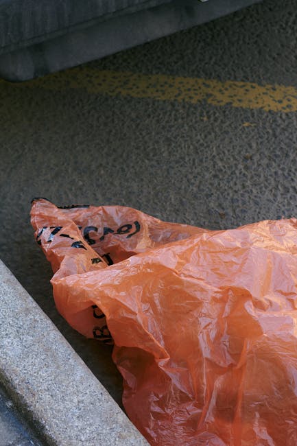 A crumpled orange plastic rubbish bag lying on a textured asphalt surface adjacent to a concrete curb in an urban or street environment. The bag appears to be partially transparent with visible crease