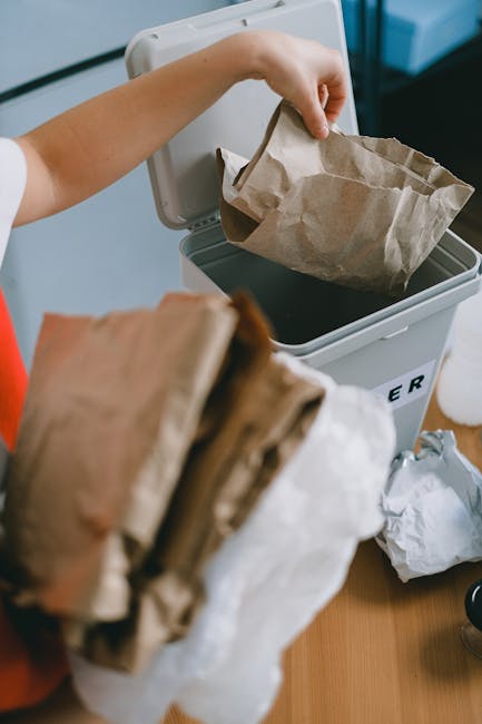 A person is shown disposing of paper waste by placing a crumpled paper bag into a grey rubbish bin with a black interior, designated for waste collection. The individual's arm is visible, wearing a sh