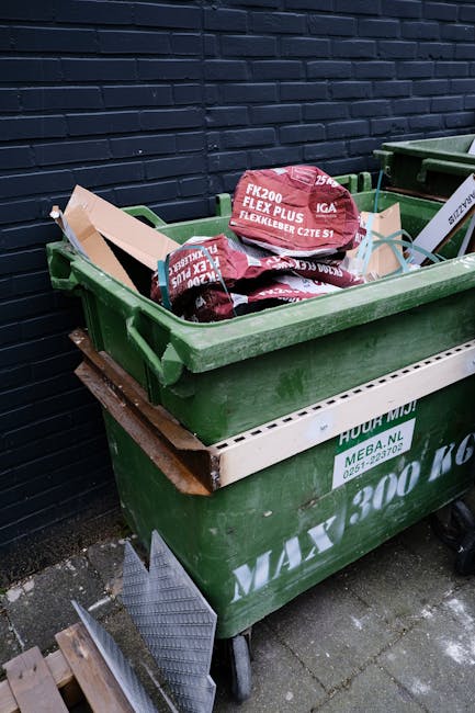 A person is shown disposing of paper waste by placing a crumpled paper bag into a grey rubbish bin with a black interior, designated for waste collection. The individual's arm is visible, wearing a sh
