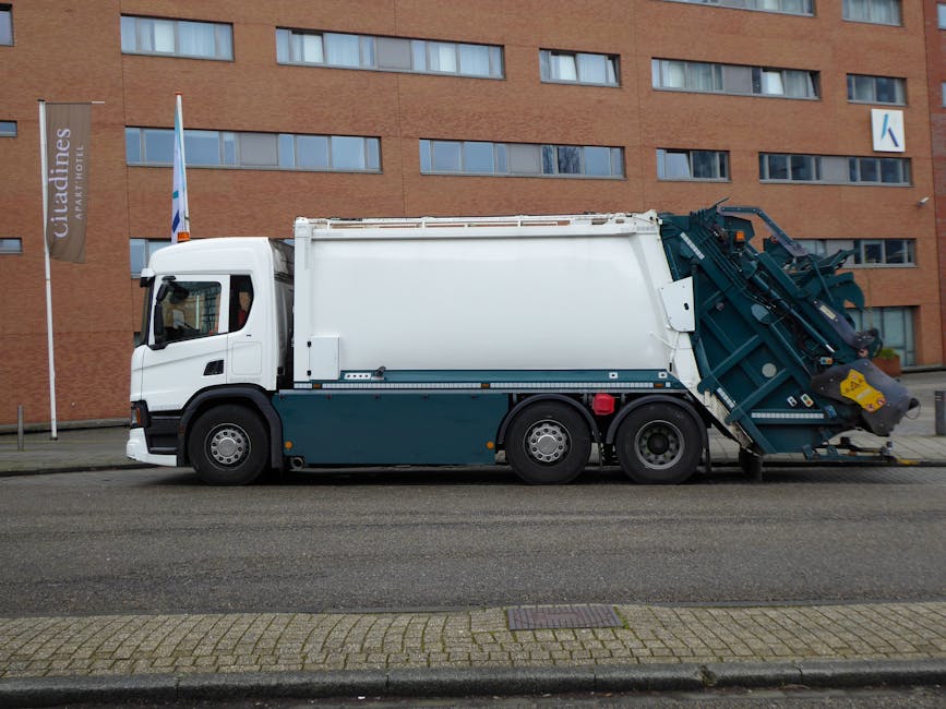 A waste collection worker wearing a high-visibility orange and grey uniform stands outdoors during dusk, next to a large yellow and white rubbish collection vehicle. The worker is positioned on the dr