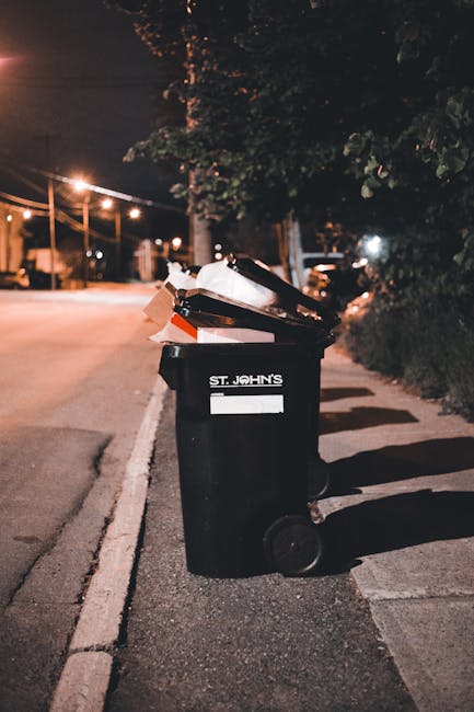 A black wheelie bin positioned on a sidewalk at night, with the label 'St. John's' visible on the front. The bin is open and filled with various types of rubbish, including cardboard, plastic packagin