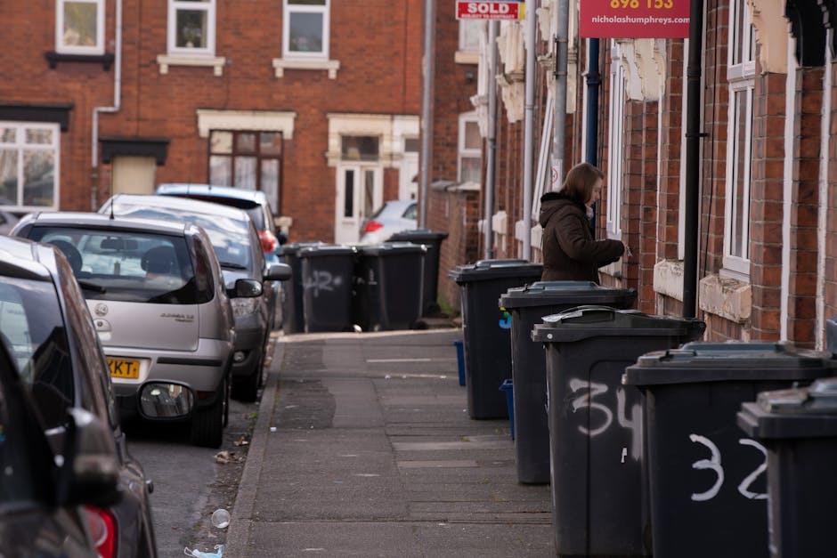 A black wheelie bin positioned on a sidewalk at night, with the label 'St. John's' visible on the front. The bin is open and filled with various types of rubbish, including cardboard, plastic packagin